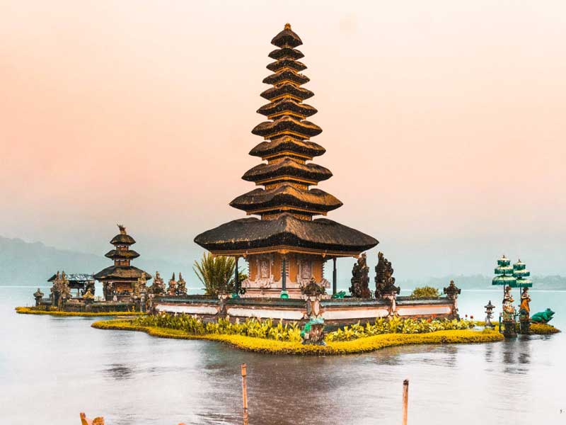 Balinese temple on a lake, with tiered roof structure. Soft pink and orange sky.