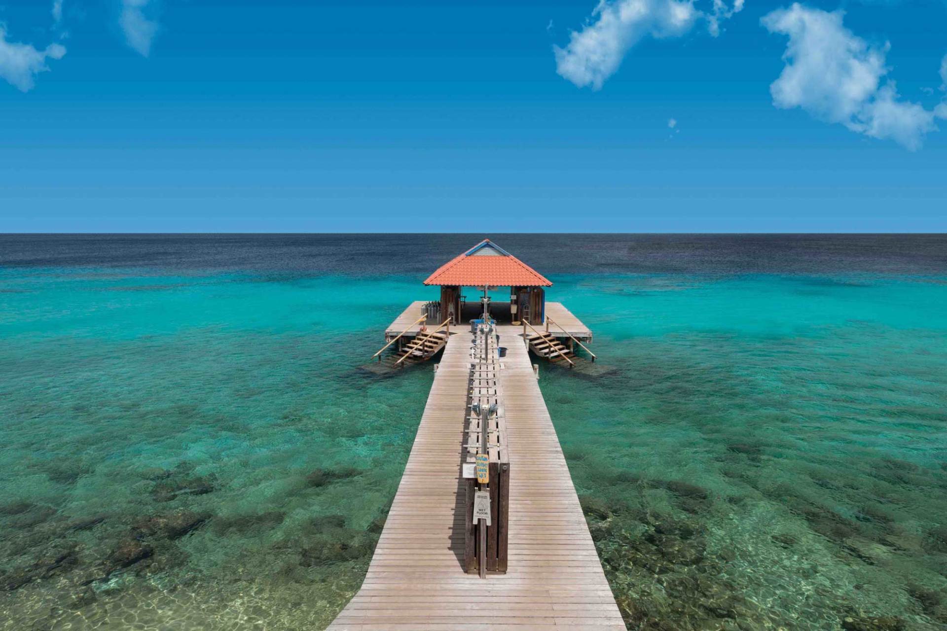 Wooden pier extending over clear turquoise water towards a gazebo under a blue sky.