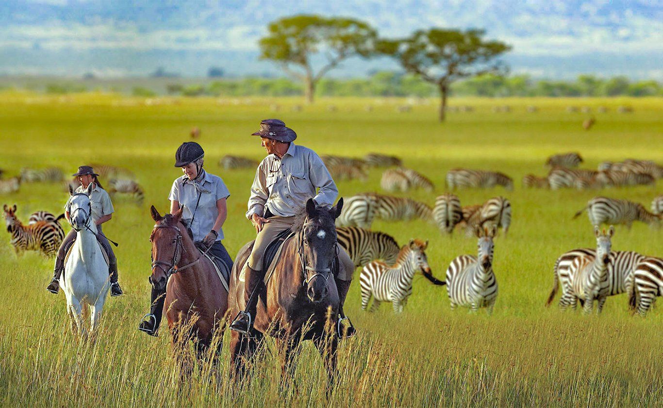People on horseback ride through a grassy plain alongside zebras.
