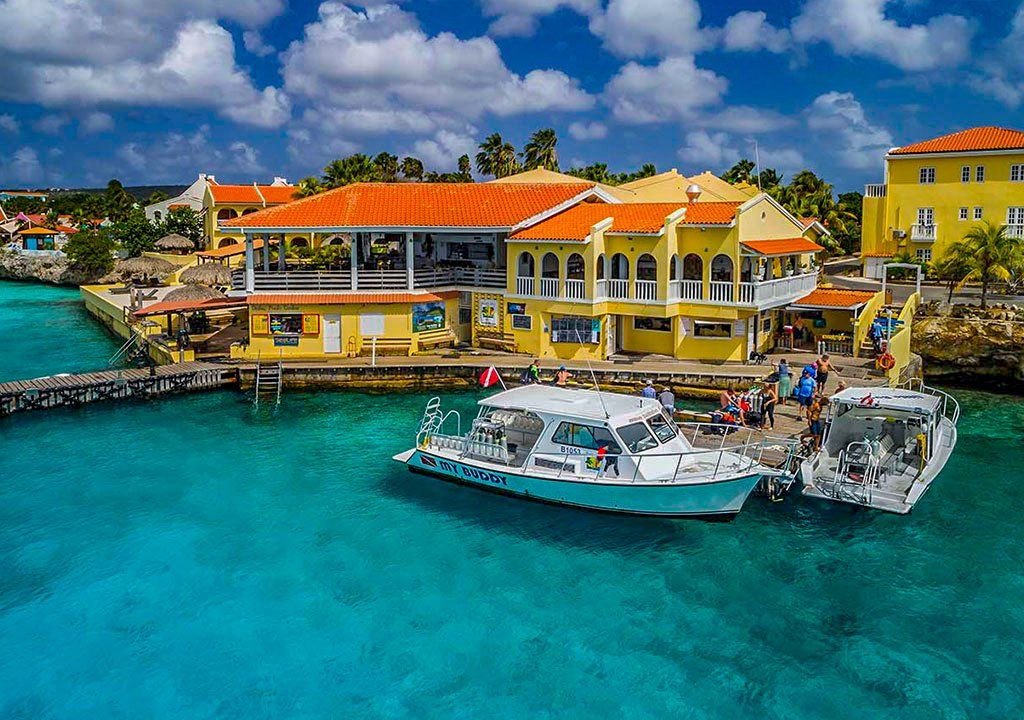 Boats docked at yellow building with orange roof on turquoise water. People stand on pier. Sunny day.