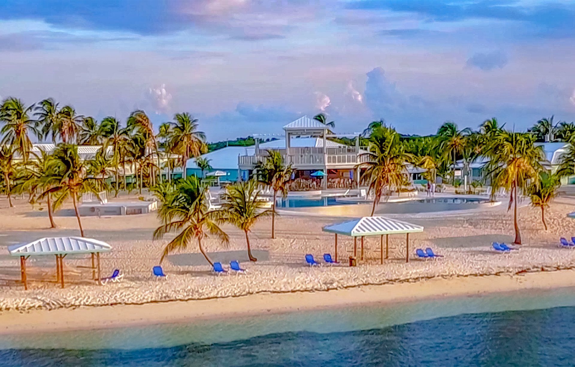 Beach with white sand, palm trees, blue chairs, and a white gazebo with a two-story building in the background.