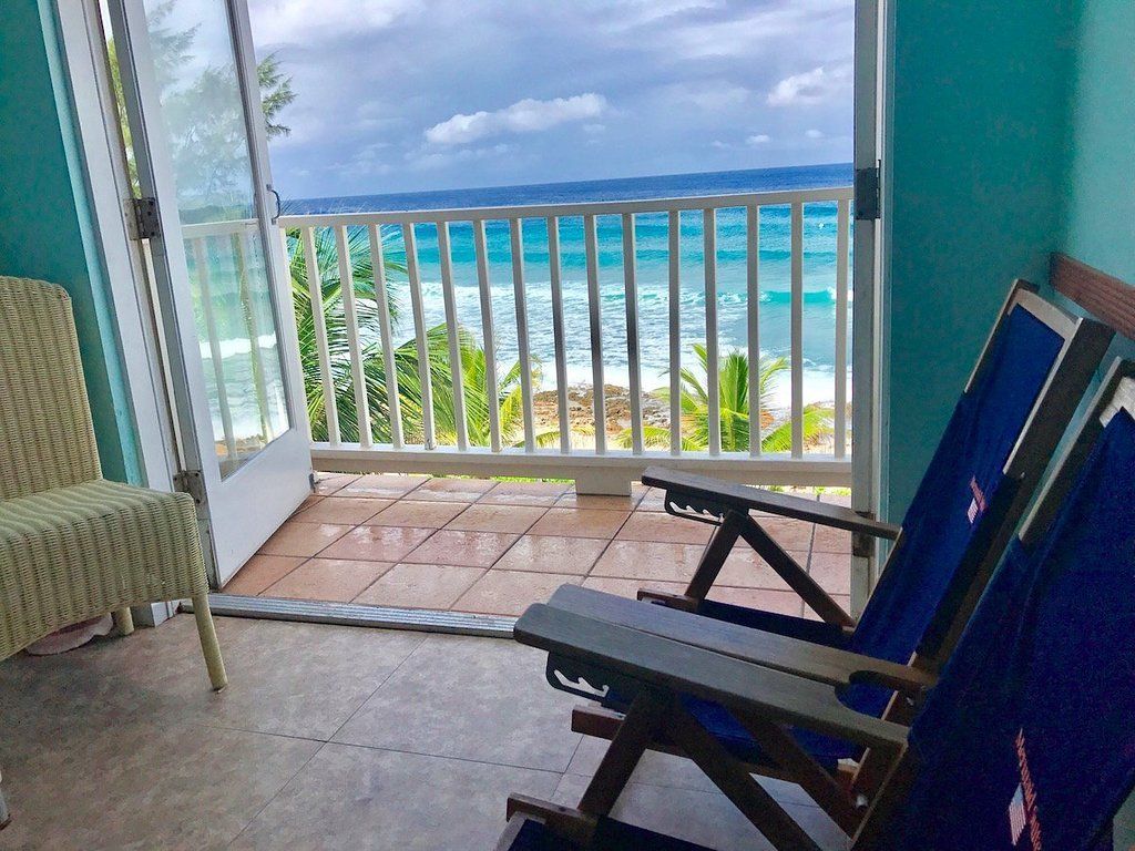 Balcony with ocean view: chairs, open door, turquoise walls, white railing, blue water, cloudy sky.