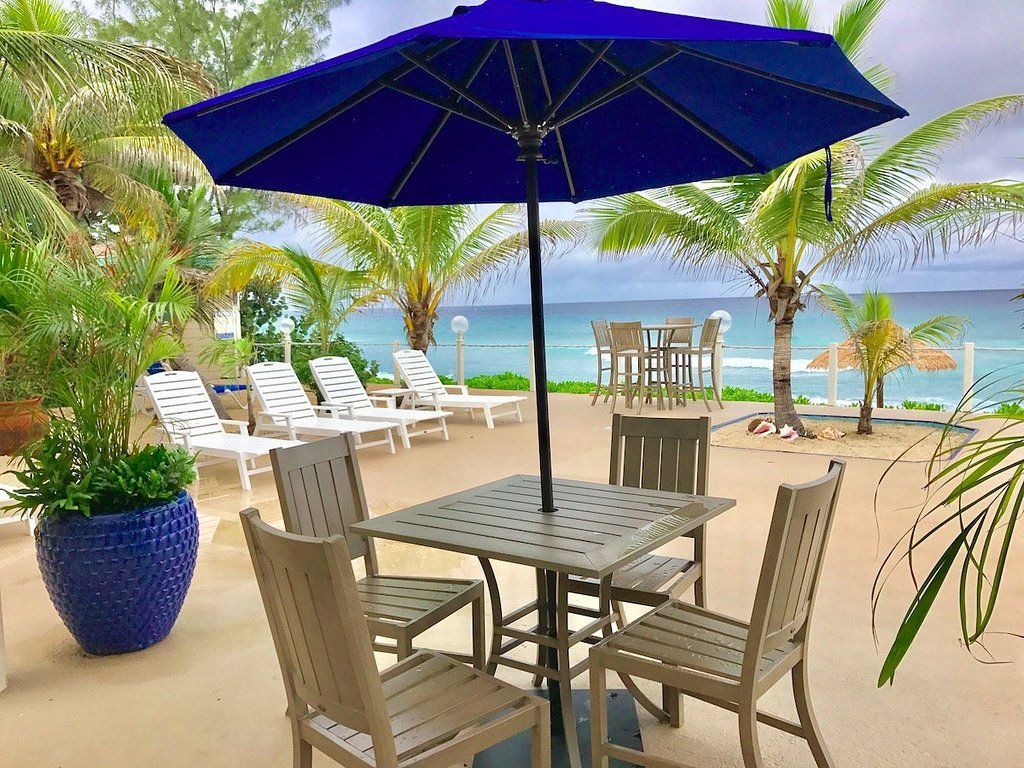 Outdoor patio with a table and chairs under a blue umbrella, with beach and ocean in the background.