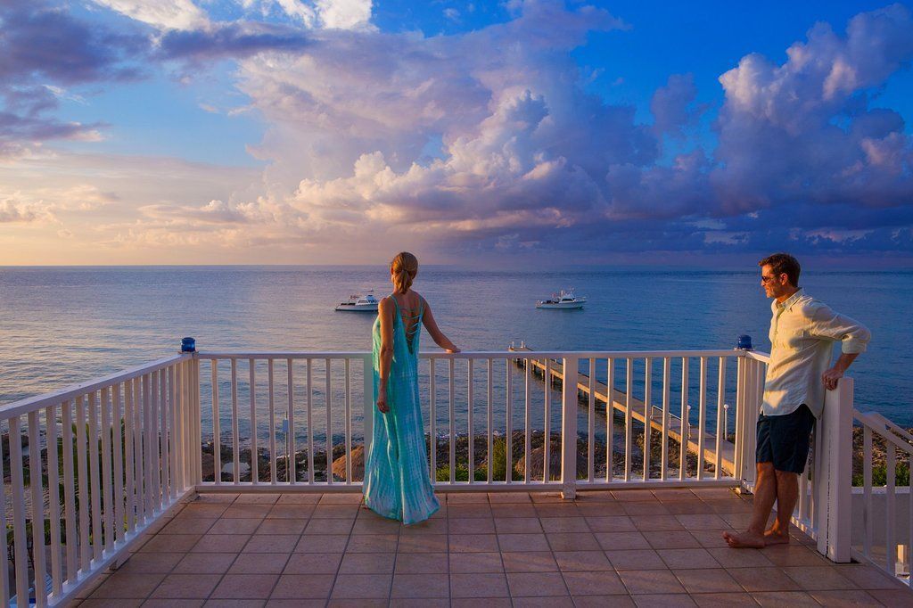 Couple on a balcony overlooking the ocean at sunset; woman in teal dress, man in shorts.
