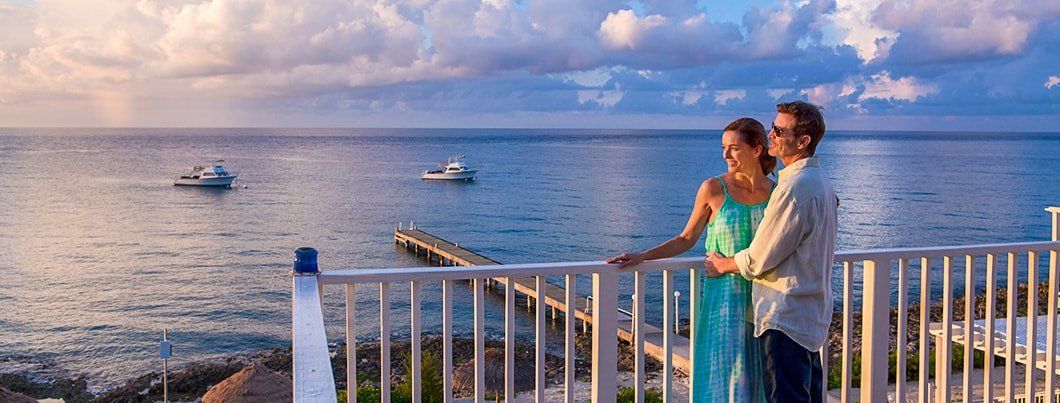 Couple on balcony overlooking ocean, boats, and pier at sunset.