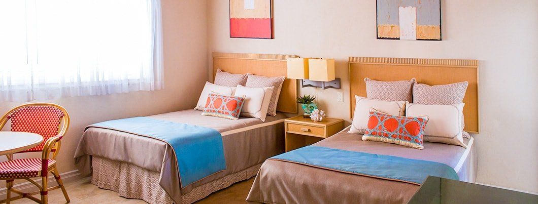 Bedroom with two twin beds, bedside table, wicker chair, and window. Blue and orange accents.