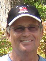 Man wearing a black hat with a white brim, glasses, and smiling outdoors.