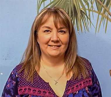 Woman with blonde-streaked hair, smiling, wearing a patterned top, in front of a blue wall with a palm tree.