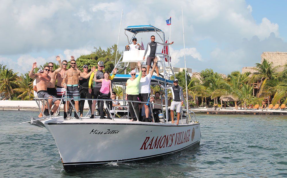 Group of people on a boat, waving and smiling in front of a beach setting.