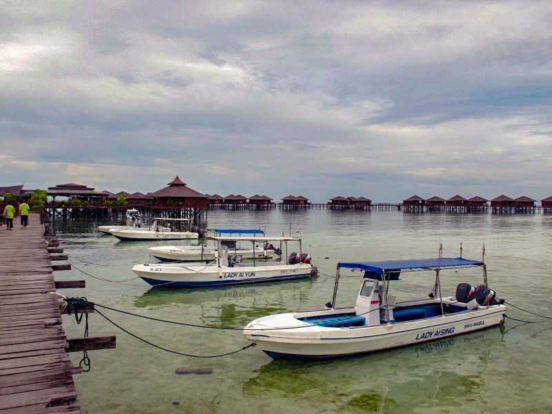 Boats docked near overwater bungalows; cloudy sky above tropical setting.