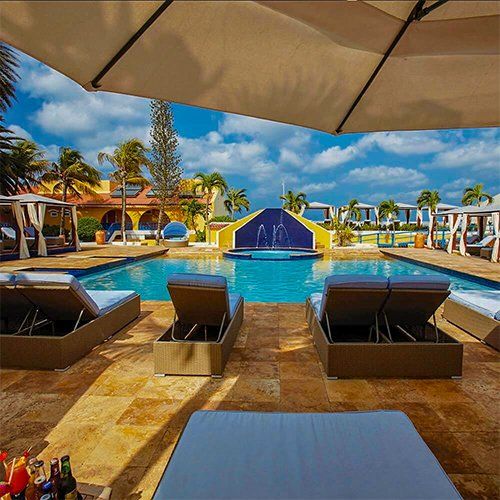Poolside scene with lounge chairs, umbrella, pool, and buildings under a sunny sky.