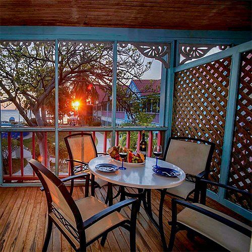 Outdoor dining area with a sunset view. Table with food and wine, four chairs, screen enclosure, wooden deck.