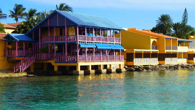 Colorful waterfront buildings with blue, pink, and yellow facades sit above clear turquoise water under a sunny sky.
