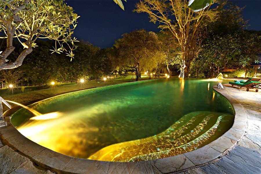 Night view of a lagoon-shaped pool, illuminated by warm lights, surrounded by trees.