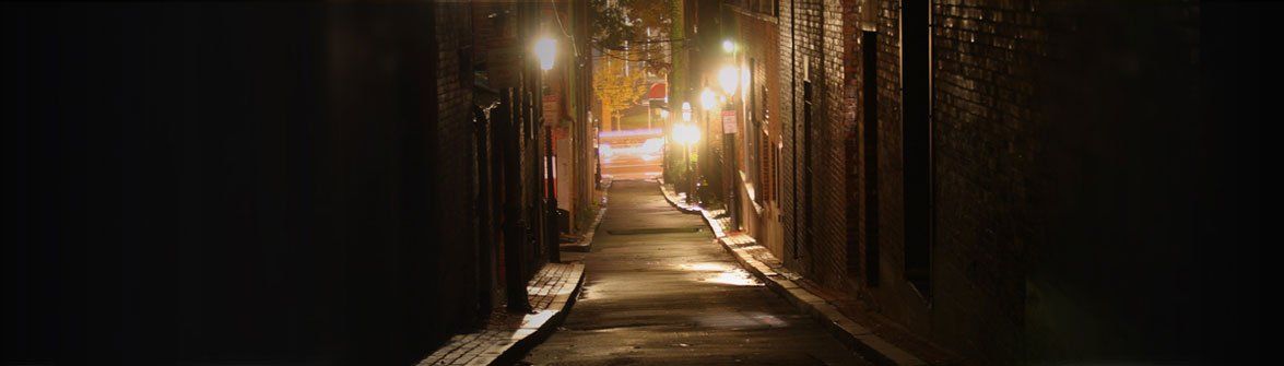 Narrow, dark alleyway at night, lit by streetlights. Wet pavement reflects the light.