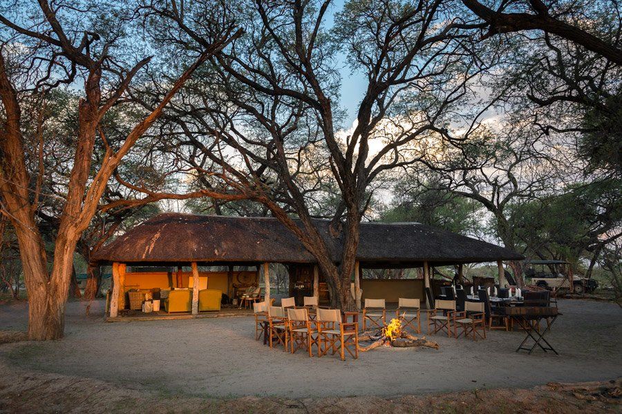 Safari camp at sunset with thatch roof, chairs, fire pit, and trees.