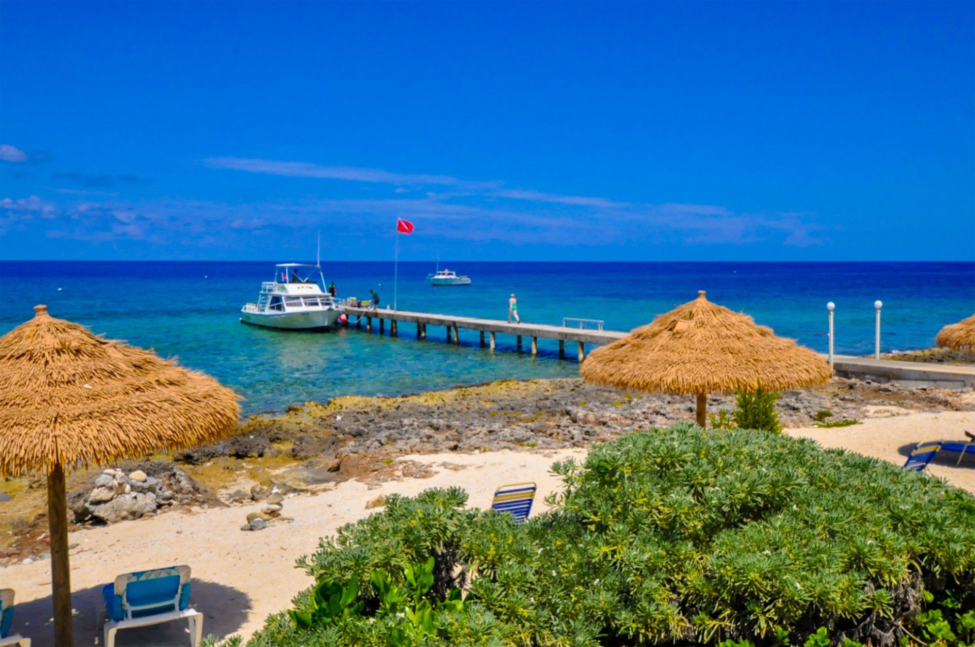 Beach scene with blue sky, ocean, a dock, boat, thatched umbrellas, and shoreline vegetation.
