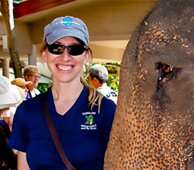 Woman smiling next to an elephant at an outdoor sanctuary; she's wearing a blue shirt and cap.