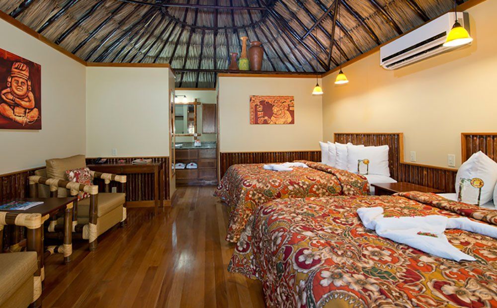 Hotel room with two beds, wooden floors, thatched ceiling, sofa, and artwork.