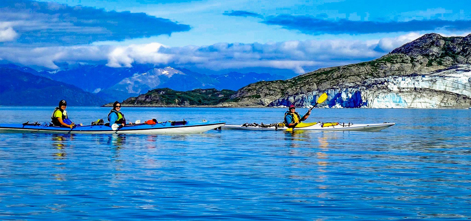 Kayakers paddling on blue water near a glacier and mountains under a blue sky.