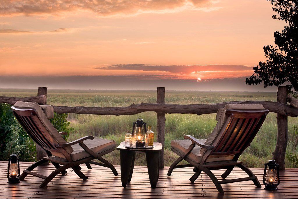 Two wooden chairs on a deck, overlooking a savanna at sunset, with lanterns and a small table.
