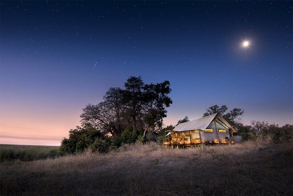 Campsite under starry night sky, illuminated with warm interior lights. Crescent moon glows.