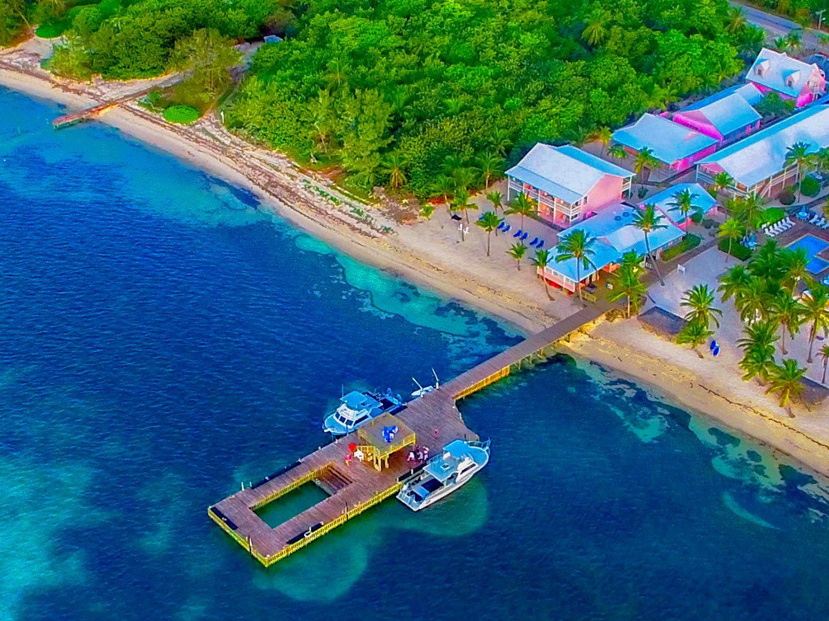 Aerial view of a dock extending into turquoise water with boats, beach, and colorful buildings, surrounded by lush green trees.