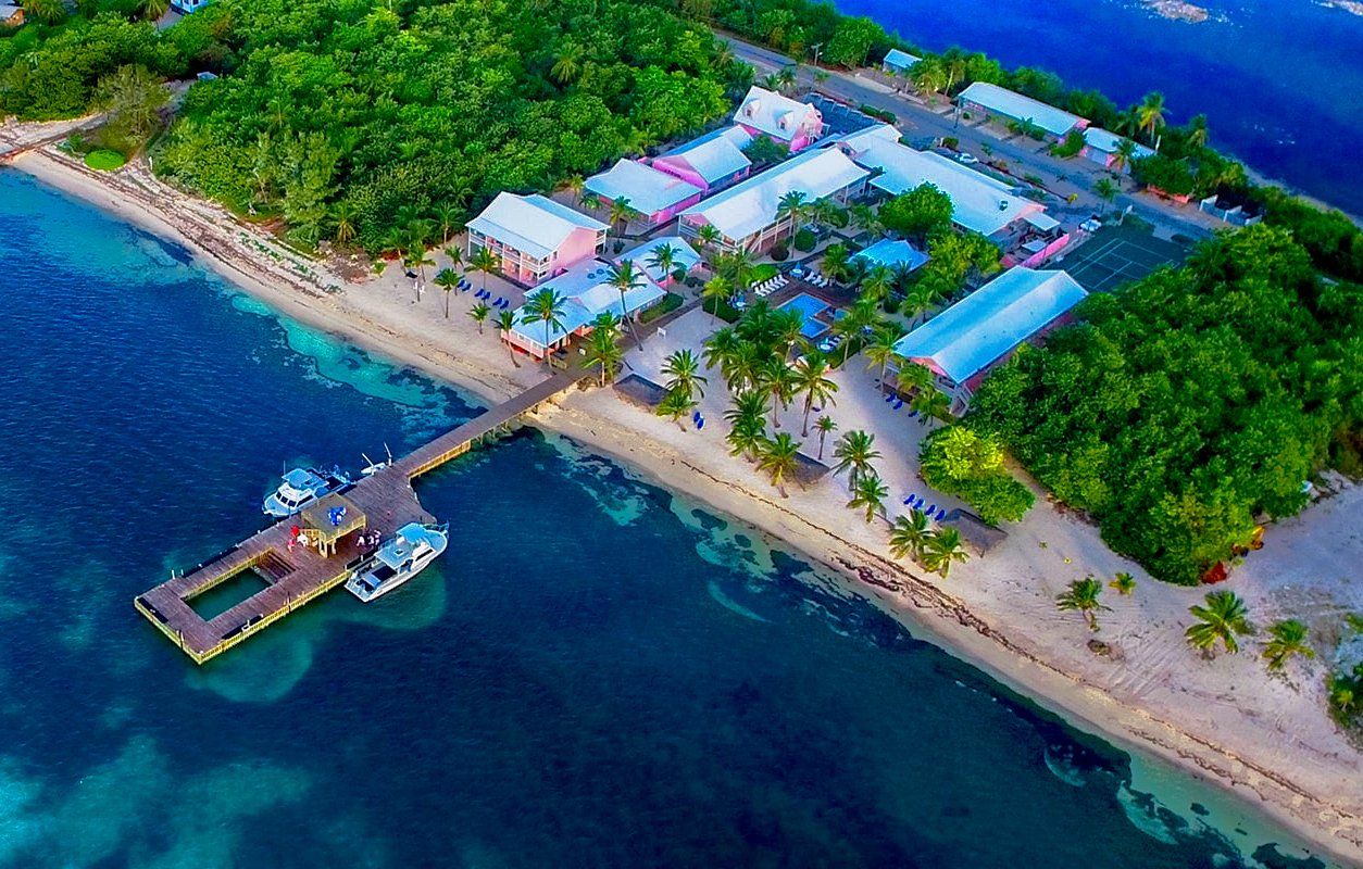 Aerial view of island resort with pier, docked boats, white sand beach, and lush green foliage.