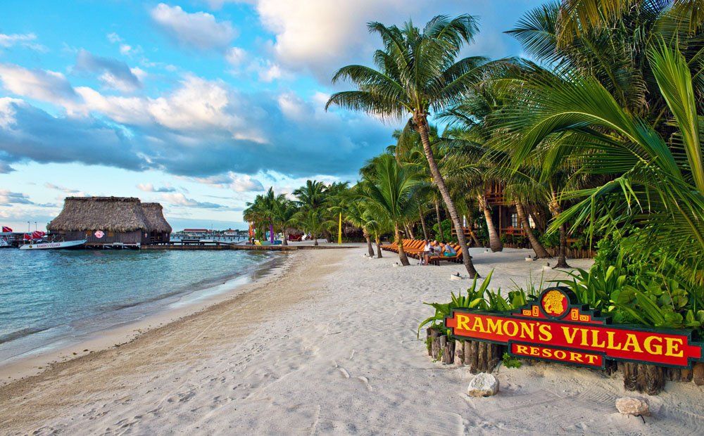 Sandy beach with palm trees and ocean, Ramon's Village Resort sign, blue sky, and a thatched-roof structure.