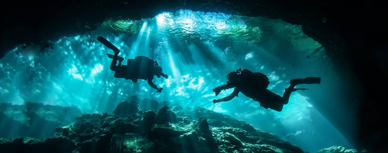 Two divers explore an underwater cave with sunbeams filtering through the water.