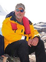 Man in yellow jacket and life vest, seated outdoors, smiles. A small bird sits nearby.