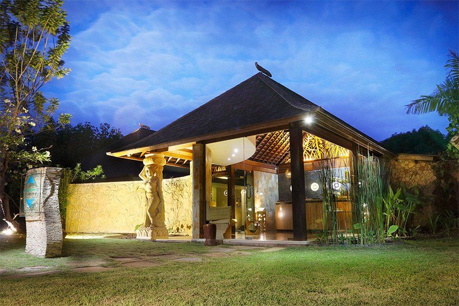 Balinese style open-air building with a thatched roof, lit at night. Stone walls, greenery and a grassy lawn surround.
