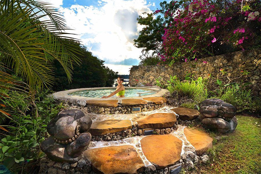 Person relaxing in a stone hot tub surrounded by greenery and flowers. Water cascades down steps.