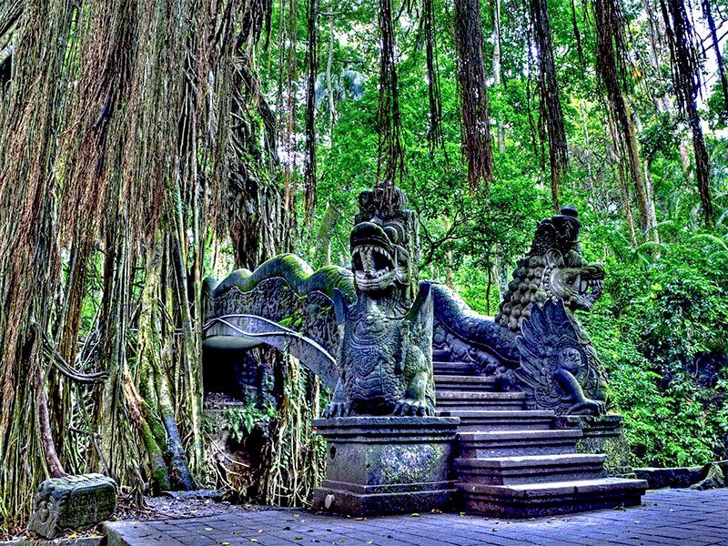 Stone statues flank a staircase in a lush forest, with hanging vines and greenery.