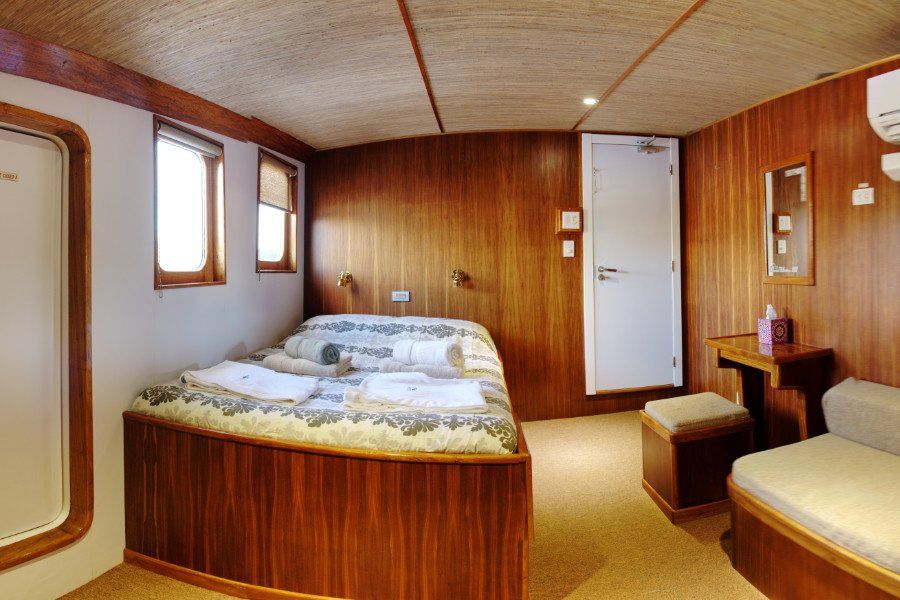 Wooden cabin on a boat with a double bed, small couch, desk, and closed door.
