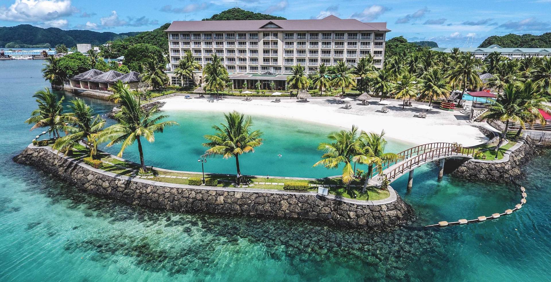 Island resort with a large hotel, sandy beach, palm trees, and clear turquoise water.