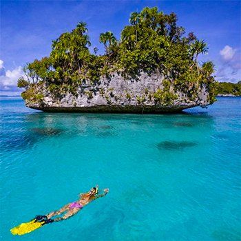 Person snorkeling in turquoise water towards a small island with lush greenery under a bright blue sky.