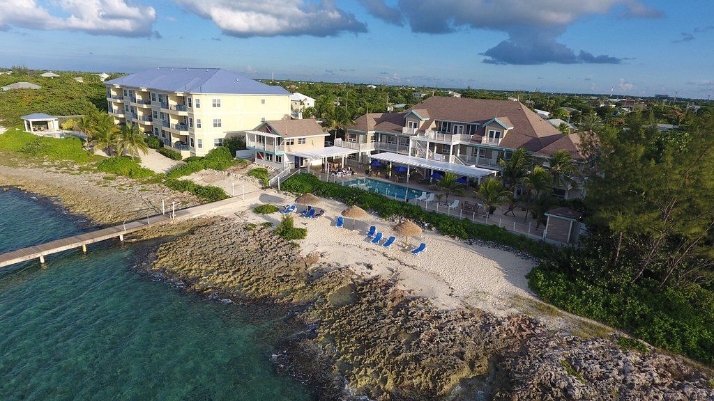Coastal resort with buildings, a small beach, a pool, and a pier, next to turquoise water under a blue sky.