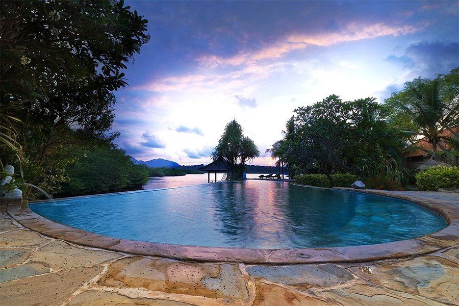 Infinity pool overlooking water, trees, and sunset. Blue, pink, and purple sky. Stone patio edge.