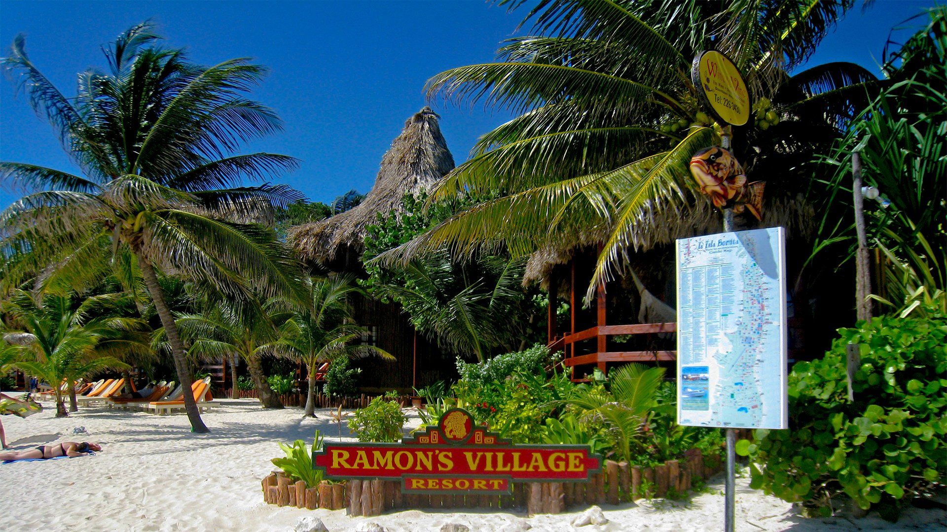Ramon's Village resort entrance with sign, thatched roof buildings, palm trees, and a white sand beach.