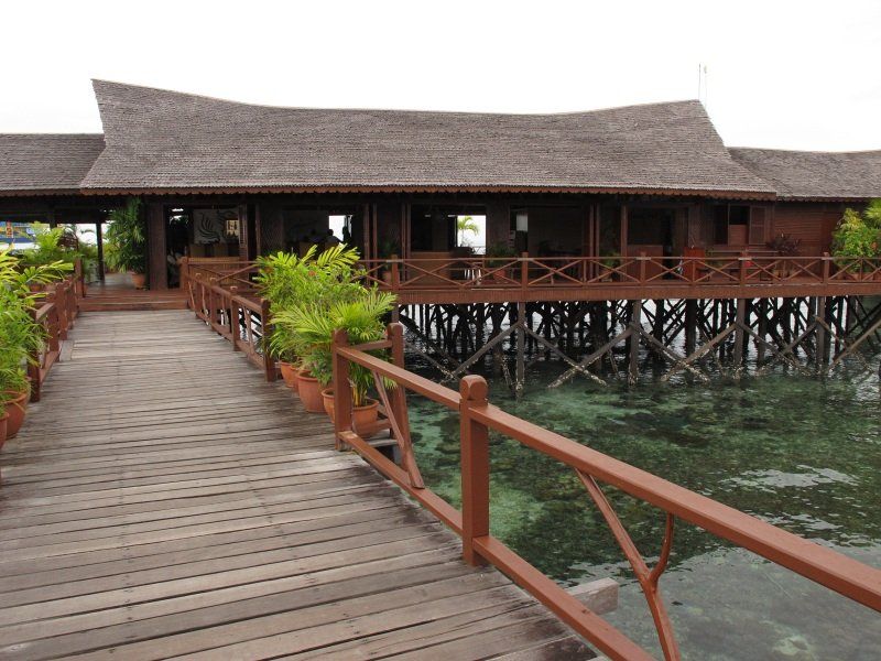 Wooden pier leading to a stilt house with a brown roof over clear water.