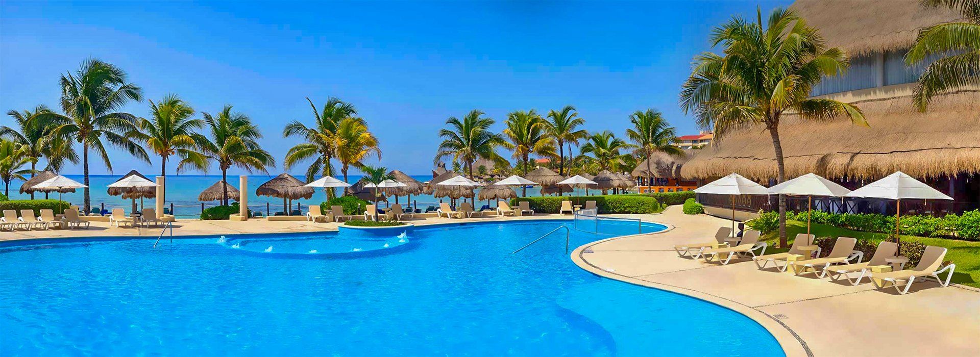 Poolside view of beach resort: blue water, palm trees, thatched-roof structures, umbrellas, and lounge chairs.