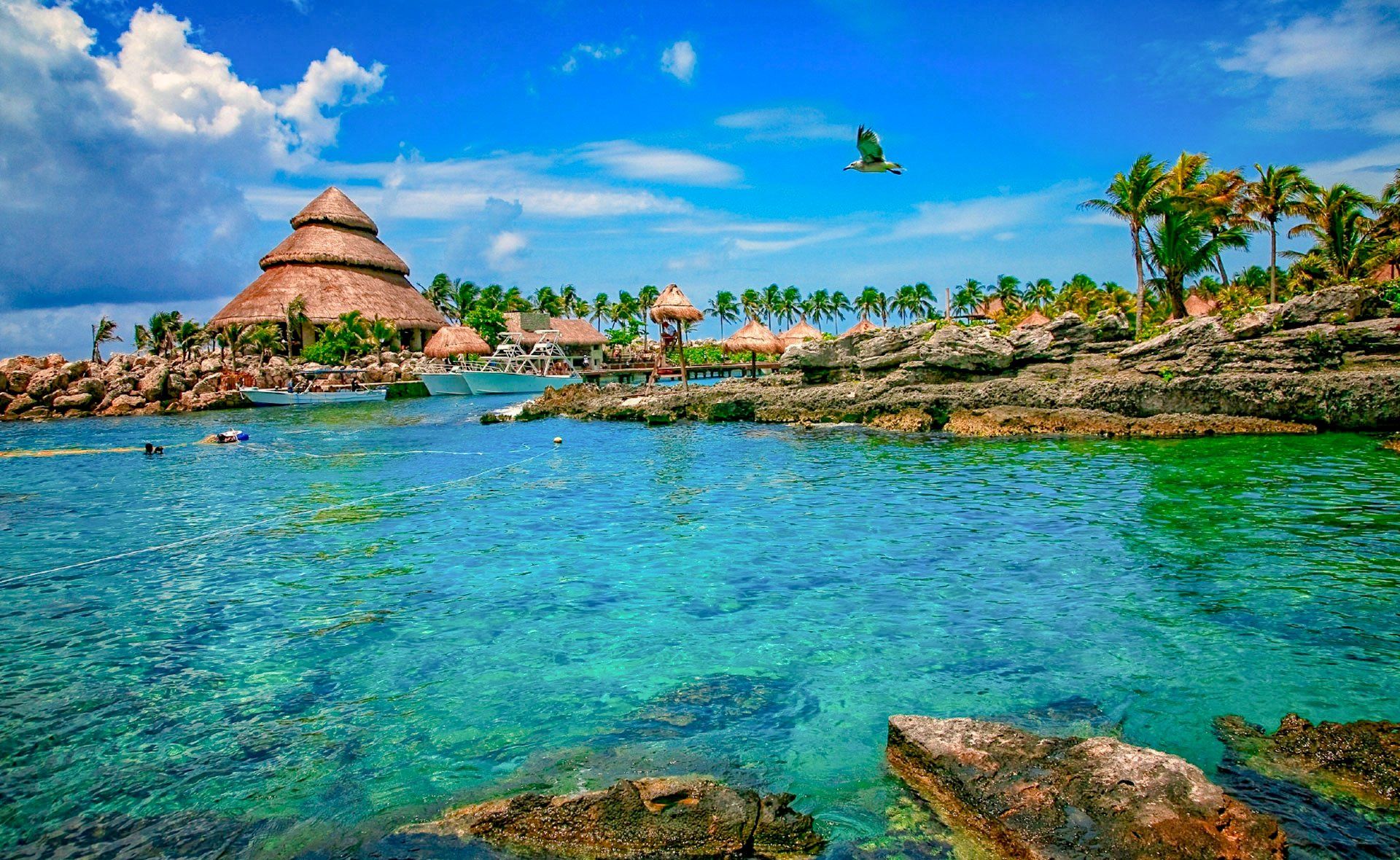 Turquoise water with rocky shore, thatched roof structures, and tropical vegetation under a blue sky.
