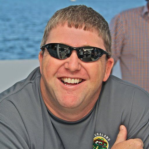 Man wearing sunglasses, smiling broadly, gray shirt, outdoors near water.