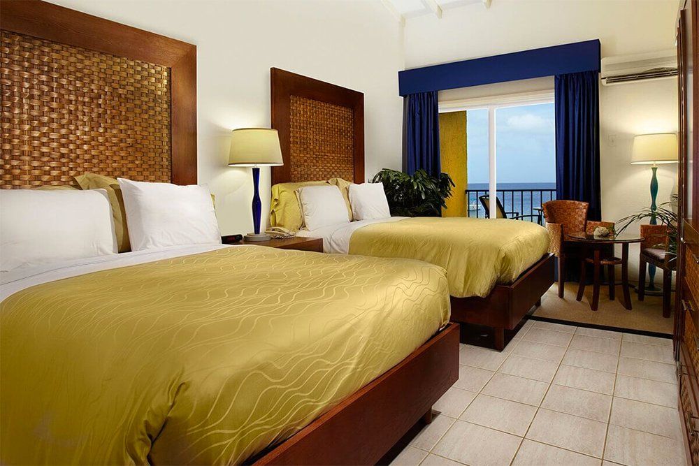 Two beds in a hotel room with woven headboards, yellow bedding, and a balcony with an ocean view.