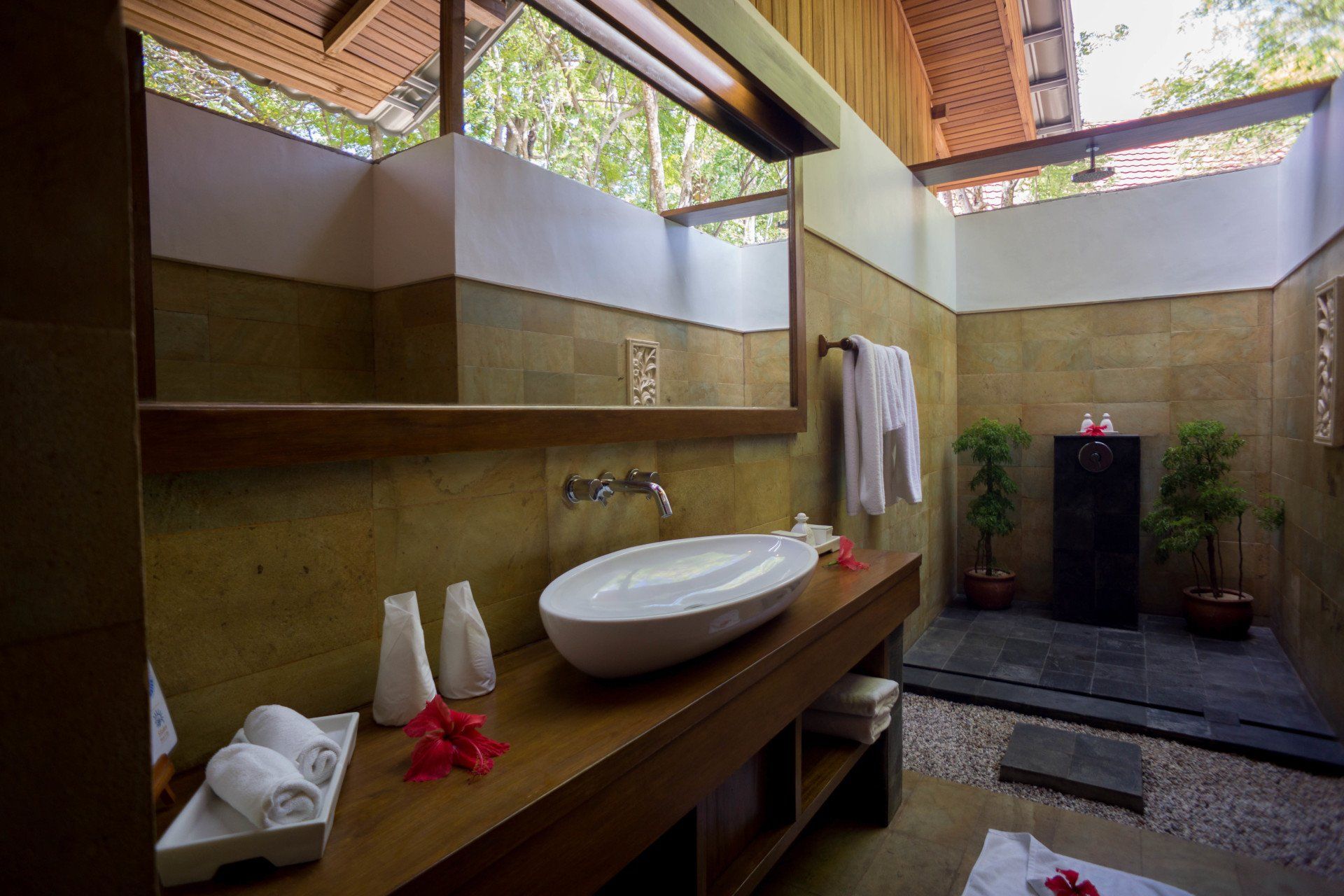 Open-air bathroom with a white sink, wooden counter, and stone walls. Towels hang; plants add greenery.