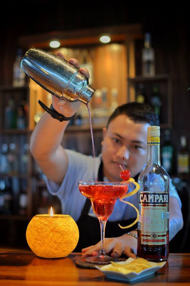 Bartender pouring a red cocktail from a shaker into a glass, next to a Campari bottle and a candle.
