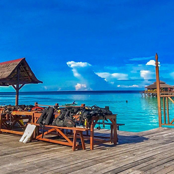 Wooden pier with scuba gear, turquoise water, and structures under a vibrant blue sky.