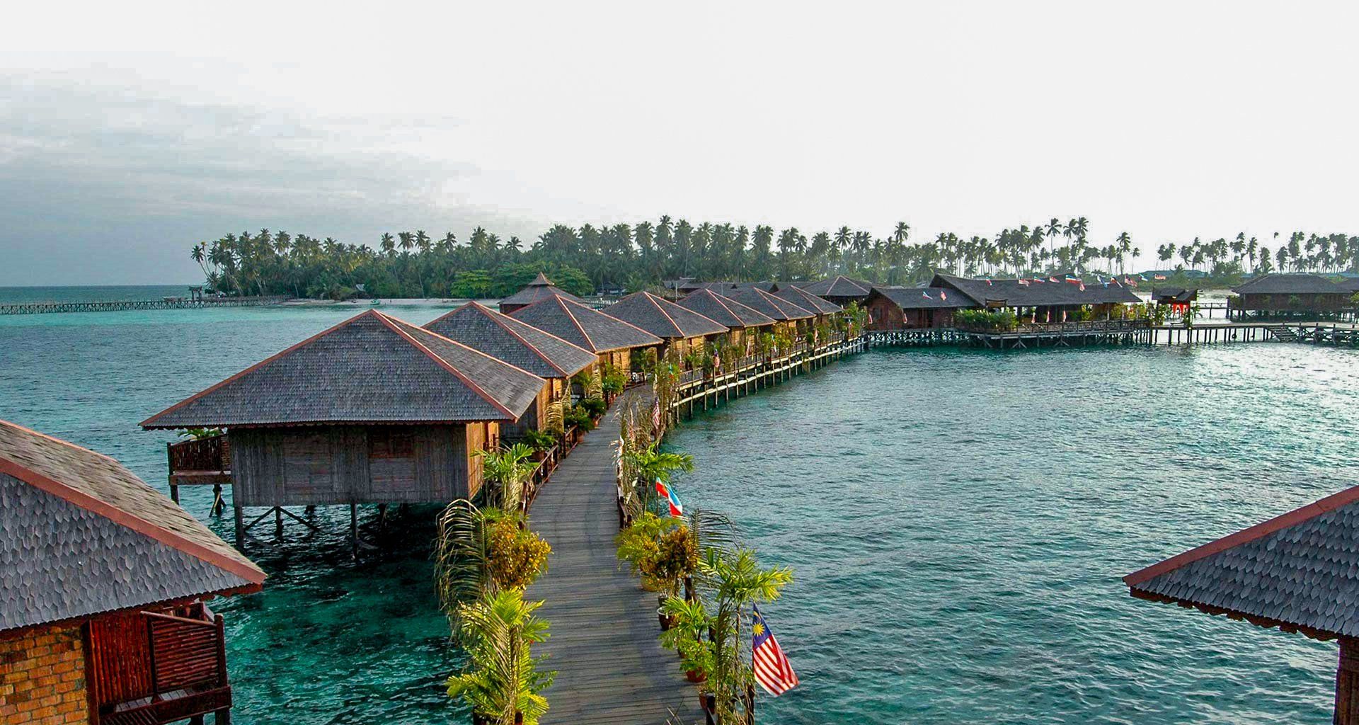 Overwater bungalows with thatched roofs connected by a wooden walkway in turquoise water.