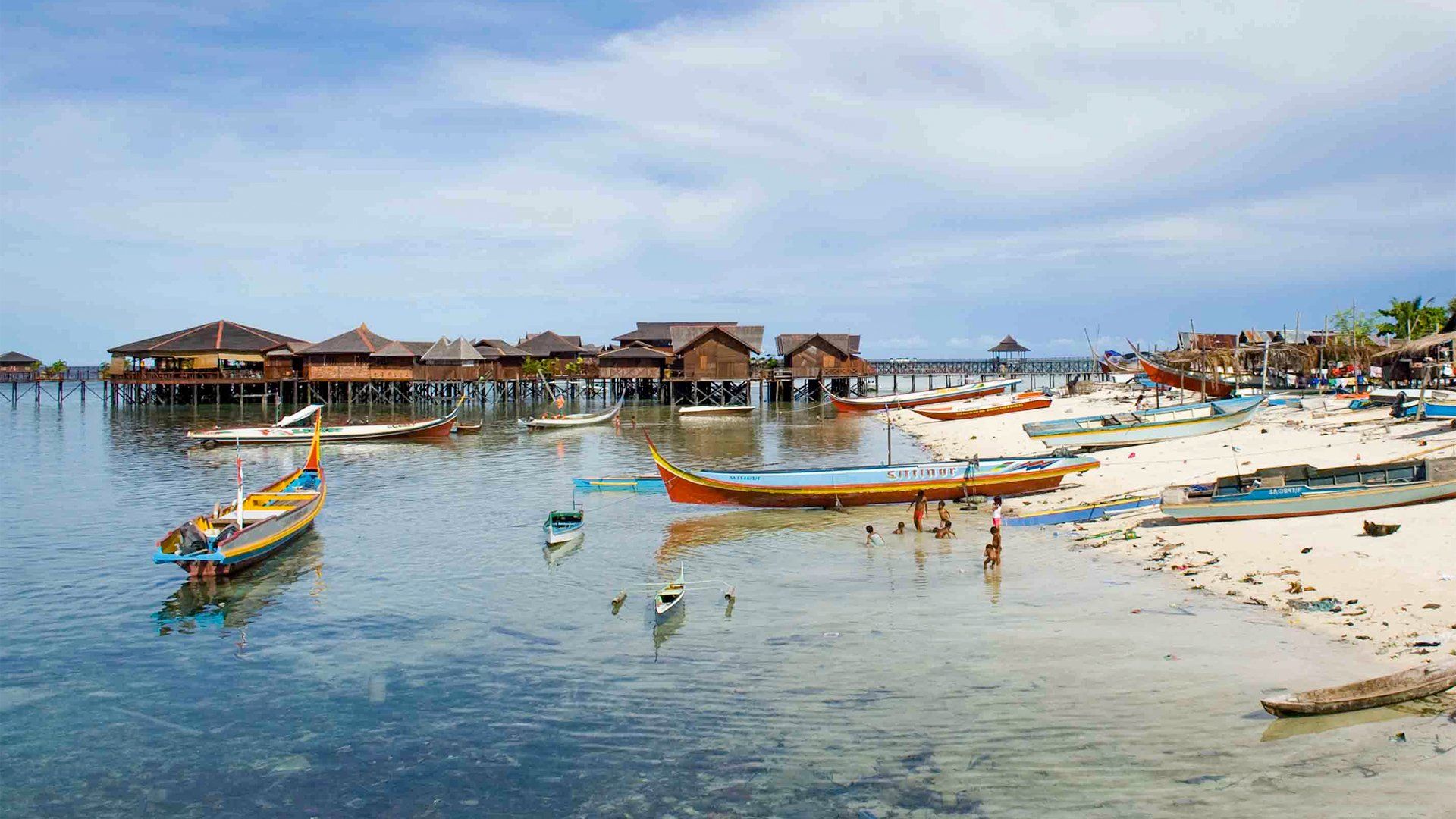 Boats dot the clear water near a sandy beach with wooden houses on stilts under a cloudy sky.